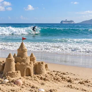 A beach scene with a sandcastle in the foreground, a surfer 