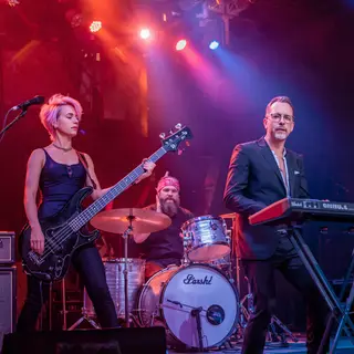 Three musicians on stage: a tall woman with short pink hair 