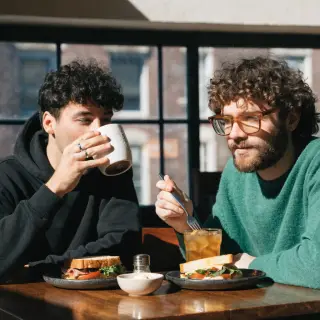 Two men at a cafe: one with a beard and glasses wearing a gr